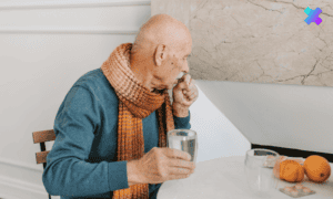 Osoba starsza kaszląca i trzymająca szklankę z wodą, objawy zapalenia oskrzeli.